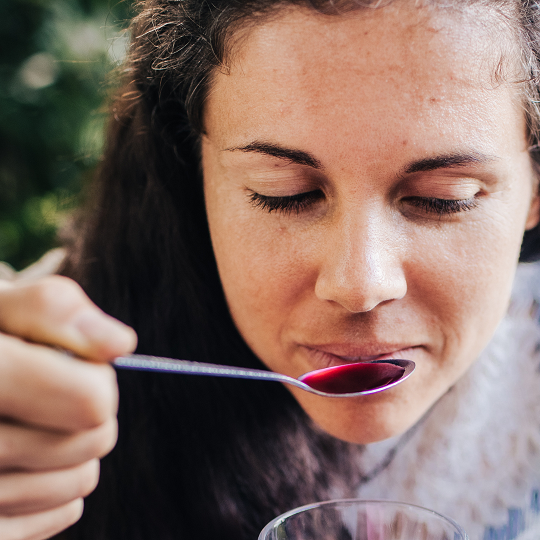 Woman up close about to take a sip of elderberry syrup off a spoon. How to use elderberry syrup effectively to stop a cold or flu. Everyday Empowered - learn herbal medicine for home herbalists.