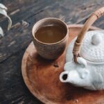 Image of ceramic mug of herbal tea next to white teapot with natural handle, on a wooden plate. Herbal Tastes