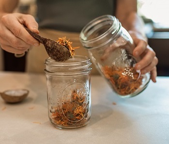 Image of putting calendula in jar with wooden spoon. How to practice herbalism on a budget. Everyday Empowered. community herbalism