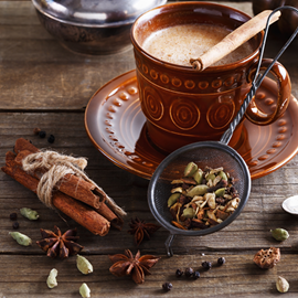 image of chai tea in brown mug with chai spices around it on the wooden bench and in the strainer. Learn to make your own homemade chai with this herbal chai tea recipe